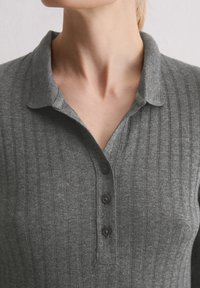 Zbliżenie na osobę w szarej, żebrowanej koszulce z odwróconym kołnierzykiem i trzema czarnymi guzikami z przodu.