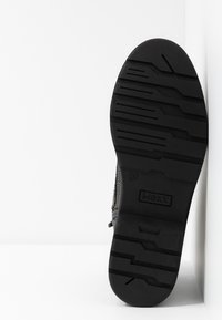 Semelle de botte noire avec des rainures profondes et le nom de la marque "MEXX" embossé, taille 37, positionnée contre un fond d'angle blanc.