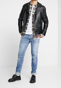 Černá kožená moto bunda přes károvanou flanelovou košili, bílé tričko, světle modré džíny a černé boty s kulatou špičkou.