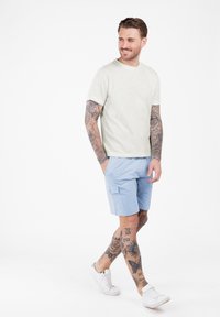 Lichtgrijs T-shirt met korte mouwen en lichtblauwe shorts met zijzakken. Het model draagt witte sneakers en heeft zichtbare zwarte tatoeages op zijn armen en benen.
