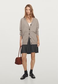 Ljust grått stickat kofta med knäppning, kombinerat med en svart plisserad kjol. Tillbehören inkluderar en brun handväska och svarta skor med strumpor.