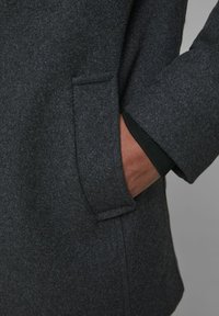 Cappotto di lana grigio con una texture sottile, dotato di una tasca laterale e un dettaglio di polsino interno nero. Focalizzato su linee pulite e design classico.