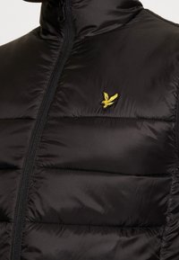 Primo piano di un giubbotto a vento nero con colletto alto, caratterizzato da un logo di un uccello giallo sull'area sinistra del petto e una zip verticale.