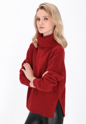 Red knitted sweater with a high ribbed collar, loose fit, and side slits, made from a textured fabric. Styled with black leggings.