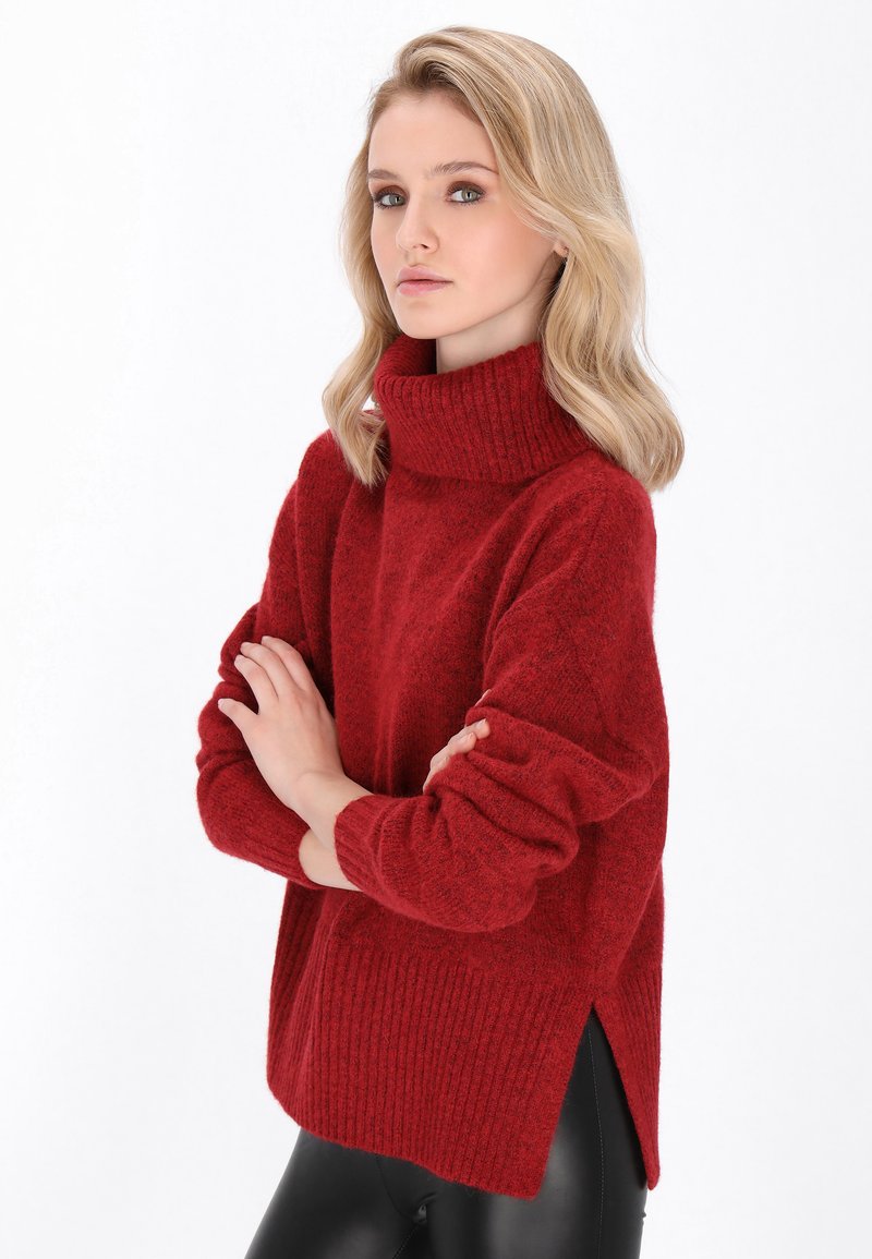 Roter Strickpullover mit hohem Rippkragen, lockerer Passform und seitlichen Schlitzen, aus einem strukturierten Stoff gefertigt. Kombiniert mit schwarzen Leggings.