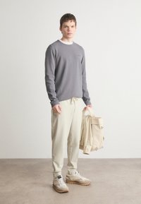 Sweatshirt gris à manches longues, pantalons beiges à cordon de serrage et baskets beiges. Modèle tenant un sac beige clair. Texture lisse, coupe décontractée.