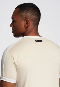 Béžové tričko s bílými rukávy, kulatým výstřihem a malou černou štítkem na zadní straně. Hladká textura a volný střih.