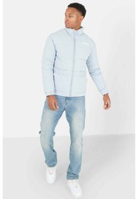 Giacca puffer azzurra chiara con zip completa e colletto alto, abbinata a jeans blu logori e sneakers bianche, in piedi su uno sfondo neutro.