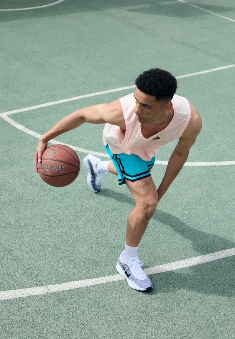 Un athlète en débardeur rose clair et en shorts turquoise dribble un ballon de basket sur un terrain vert, portant des chaussures de sport blanches et grises.