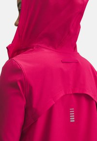 Lys pink hættejakke med en glat tekstur, der har et bagdesign i et sølvreflekterende mønster og en funktionel baglomme.