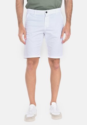Uomo con pantaloncini bianchi fino al ginocchio, maglia verde e sneakers bianche, in piedi con le braccia rilassate lungo i fianchi davanti a uno sfondo bianco.