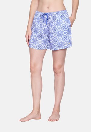 Femme portant un short à motifs floraux bleu et blanc avec un cordon de serrage, debout pieds nus devant un fond uni.