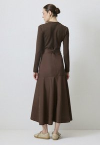 Robe longue marron à manches longues avec une taille cintrée et une jupe évasée. Le tissu semble lisse. Possède un lien au dos et un design jusqu'au sol.