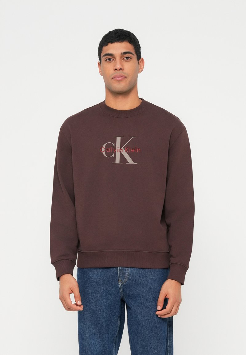 Hnědá mikina s kulatým výstřihem a žebrovanými manžetami, s výrazným světle šedým logem "cK" a červeným nápisem "Calvin Klein". Doplněná modrými džínami.