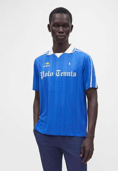 Un homme portant un polo de tennis bleu avec un col blanc, le logo US Open et un pantalon bleu marine, debout devant un fond blanc uni.