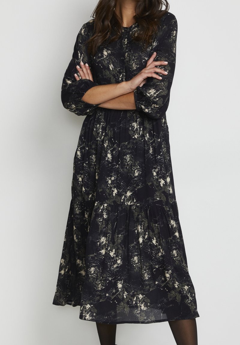 Femme portant une longue robe noire avec des motifs abstraits blancs et gris, debout les bras croisés devant un fond uni.