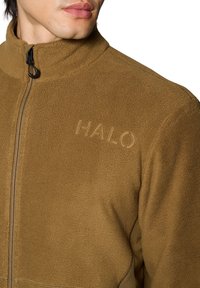 Close-up van een persoon die een bruin fleecejack draagt met een voorrits en "HALO" geborduurd op de borst.