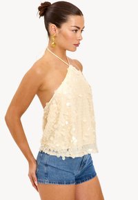 Jeune femme avec un chignon portant des boucles d'oreilles en coquillage dorées, un haut à sequins crème sans manches et un short en denim bleu, faisant face à droite sur fond blanc.