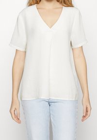 Bílá blůza s výstřihem do V a krátkými rukávy, vyrobená z lehké tkaniny. Volný střih s jemnou texturou. V kombinaci se světlými modrými džíny.