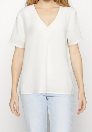 Blusa blanca de cuello en pico con mangas cortas, hecha de tela ligera. Corte holgado con una textura sutil. Combinada con jeans de color azul claro.