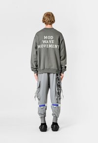 Sudadera gris con el texto "MOVIMIENTO MOD WAVE" en la parte posterior, combinada con pantalones cargo grises que tienen bolsillos laterales y acentos reflectantes.