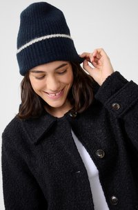 Bonnet côtelé marine avec une bande blanche ; en laine texturée. Manteau noir texturé avec de gros boutons, porté sur un haut blanc.