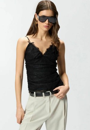 Donna che indossa un top nero in pizzo, pantaloni beige, cintura nera, orecchini a cerchio dorati e occhiali da sole neri oversize, in piedi su sfondo bianco.