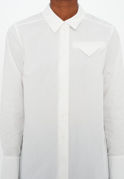 Camicia bianca con colletto a punta, maniche lunghe e una piccola tasca sul petto con una patta triangolare. Texture del tessuto liscia.
