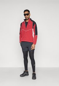 Sarkans un melns zip-up jakas ar baltajiem akcentiem, melni īsām piedurknēm, pieguļošas melnas legingi un melna sporta apavi ar Nike logo.