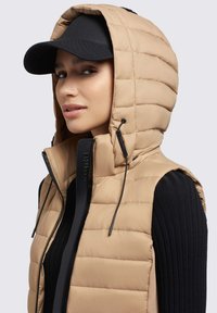 Gilet imbottito beige con collo alto e cappuccio, caratterizzato da una trapuntatura verticale e un dettaglio della cerniera nera. Indossato con un cappello nero e una maglia a costine.