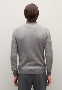 Grauer gerippter Pullover mit hohem Kragen aus weichem Stoff. Verfügt über lange Ärmel und eine taillierte Passform. Kombiniert mit grauen Hosen.
