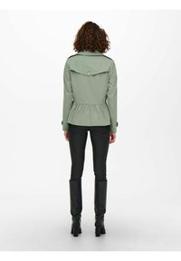 Giacca peplum verde con un'imbottitura a pulsante, maniche lunghe e vita cincta, abbinata a pantaloni neri slim-fit e stivali al ginocchio.