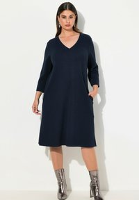 Tmavě modré pletené šaty s výstřihem do V, třičtvrtinovými rukávy, bočními kapsami a volným střihem, doplněné kovovými kotníkovými botami.