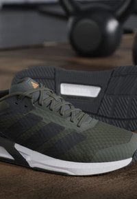 Chaussure de sport vert foncé et noire avec semelle blanche exposée sur un sol en bois, avec des kettlebells flous en arrière-plan.