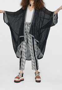 Černý průhledný kimono s širokými rukávy, vrstvené přes šedý top a kalhoty s černo-bílým vzorem, doplněné odvážnými platformovými sandály.