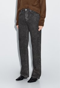 Jeans in denim nero con una finitura leggermente sbiadita, taglio dritto, design a cinque tasche e chiusura con bottone, abbinati a un maglione marrone.