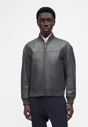 Junger Mann steht und trägt eine dunkelgraue Lederjacke mit Reißverschluss, gerippten Bündchen und Kragen, weißes Hemd und dunkle Hose vor einem schlichten Hintergrund.