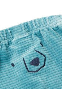 Detailní záběr na světle modrou kordurovovou látku s tmavě modře vyšitým abstraktním obličeje a elastickým páskem.