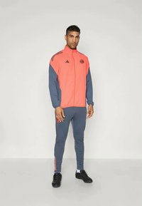 Red and grey athletic tracksuit with zip-up jacket and tapered pants. Features three black stripes and a logo on the upper left chest.
