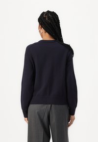 Tmavě modrý pletený svetr s dlouhými rukávy, žebrovanými manžetami a lemem, s jednoduchým kulatým výstřihem a hladkou texturou; nošený s šedými pruhovanými kalhotami.