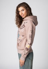 Lichtroze hoodie met een ontspannen pasvorm, voorzien van kleurrijke bedrukte ontwerpen, een trekkoordkap en geribbelde mouwen. Draagt met blauwe denim jeans.