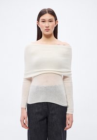 Svetložltý krémový sveter so spadnutými ramenami a dlhými rukávmi, vyrobený z jemného úpletu, ktorý sa postupne zlieva do svetlosivej lemovky. Nosený s tmavými nohavicami.