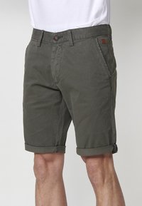 Tumši olīvzaļi vīriešu prezidenta šorti ar apakšmalu, ar priekšējiem kabatām, jostas cilpiņām, pogu aizdari, valkāti ar iekļautu baltu T-kreklu.