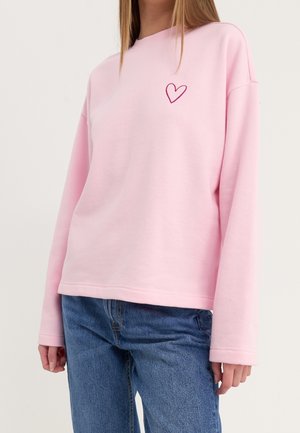 Person iført en lyserød langærmet sweatshirt med et lille mørkerødt broderet hjerte på venstre bryst og blå jeans.