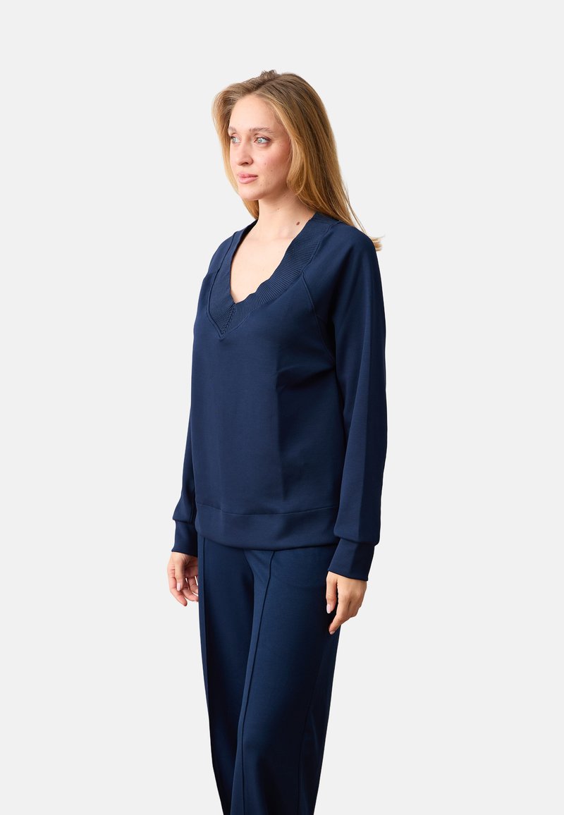 Femme aux cheveux clairs portant un haut bleu marine à manches longues et col en V, avec un pantalon assorti, debout et regardant légèrement vers la gauche.