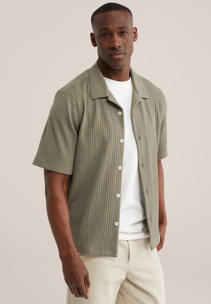 Homme portant une chemise olive côtelée à manches courtes et boutonnée par-dessus un t-shirt blanc et un pantalon beige, debout devant un fond beige clair.