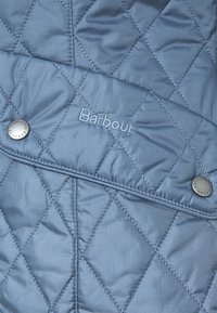 Niebieska pikowana tkanina z wzorem w romby, z naszytym logo i metalowymi zatrzaskami na wzorze. Gładka tekstura, lekki materiał.