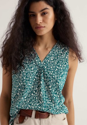 Naine pikkade lokkis juustega, kes kannab merevaigurohelist varrukateta topi valge lillemustriga ja beežeid pükse pruuni vööga, neutraalne taust.