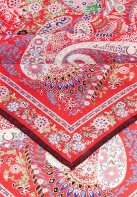 Red patterned scarf featuring floral and paisley designs in green, blue, and pink. Edged with a darker trim for contrast. Cotton material.