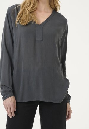 Kvinde iført en løs, mørkegrå langærmet V-halsbluse med knapmanchetter og sorte bukser mod en ensfarvet baggrund.